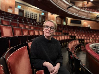 A woman is sitting in a theatre auditorium, facing towards the camera.