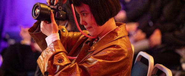 A photographer with a bob and leather jacket looks through the lens of her camera.