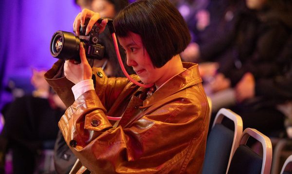 A photographer with a bob and leather jacket looks through the lens of her camera.