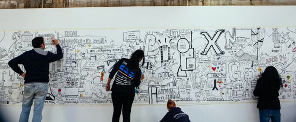 4 people stand along a white wall covered in drawings and doodles, they are adding their own art to it.