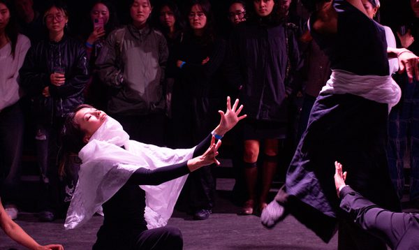 A performer stands on one-knee with white fabric wrapped around her. Another performer is twisting in the air.