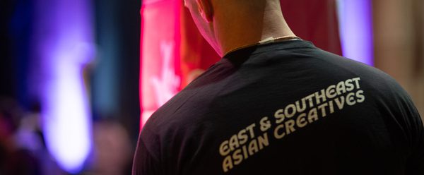 A bald crew member has his back to the camera. He is backstage and his t-shirt reads "East & South East Asian Creatives"