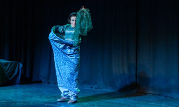 Ruxy Cantir stands on stage in front of a black curtain. They are dressed as a clown and holding a net. The lighting and costume are blue and green.
