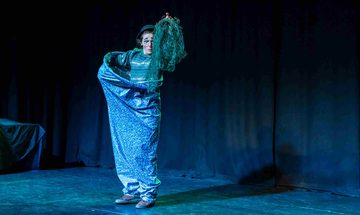 Ruxy Cantir stands on stage in front of a black curtain. They are dressed as a clown and holding a net. The lighting and costume are blue and green.