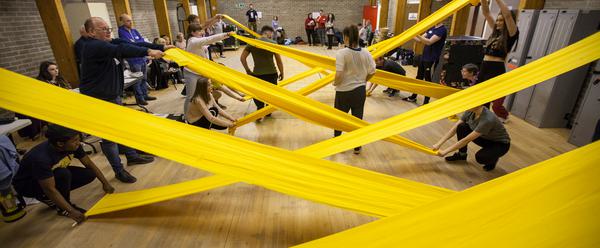 A long room with people holding rolls of yellow fabric stretched out to create a criss-cross shape along the room.