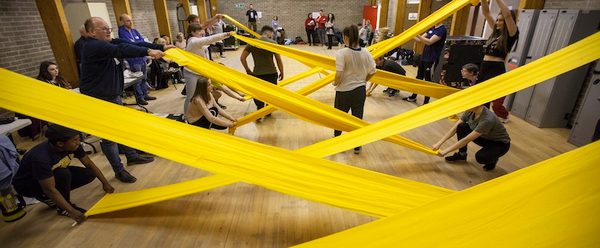 A long room with people holding rolls of yellow fabric stretched out to create a criss-cross shape along the room.
