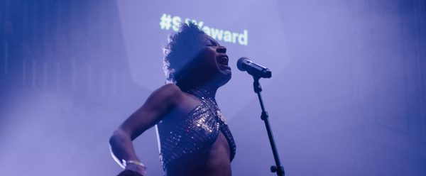 Beldina stands on-stage and sings into a microphone. The lighting is purple.