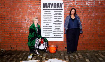 Two women are standing next to a red brick with a poster that reads MAYDAY with a list of participating artists. They have protest signs and a megaphone.