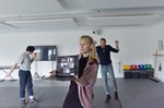 Person in a rehearsal studio holds up an open laptop showing a video call, while two others move in the background near a screen and equipment in a bright, minimal space
