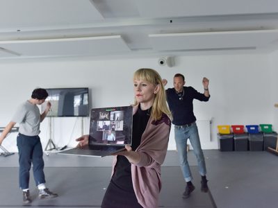 Person in a rehearsal studio holds up an open laptop showing a video call, while two others move in the background near a screen and equipment in a bright, minimal space