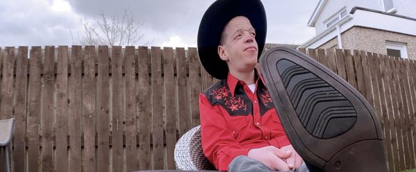 Allen McEwan sits in full rodeo get up with his cowboy booted feet propped up