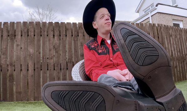 Allen McEwan sits in full rodeo get up with his cowboy booted feet propped up