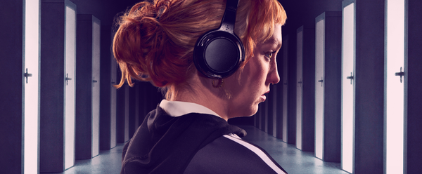 A young person with long red hair stands in a hallway, doors on either side extend int the background. She is wearing a pair of black headphones.