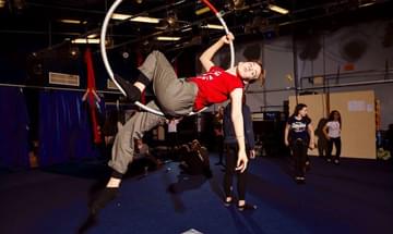A teenager with a red top is suspended on a hoop. She has one leg and one arm hanging down, the other holding the hoop.