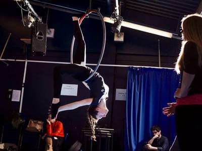 A teenager is suspended on a hoop, her head and leg facing down, her other leg up on the hoop. She is in a gym room and other are watching.