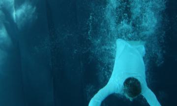 a man is plunging vertically into a pool, fully dressed with a whit shirt and black trousers.