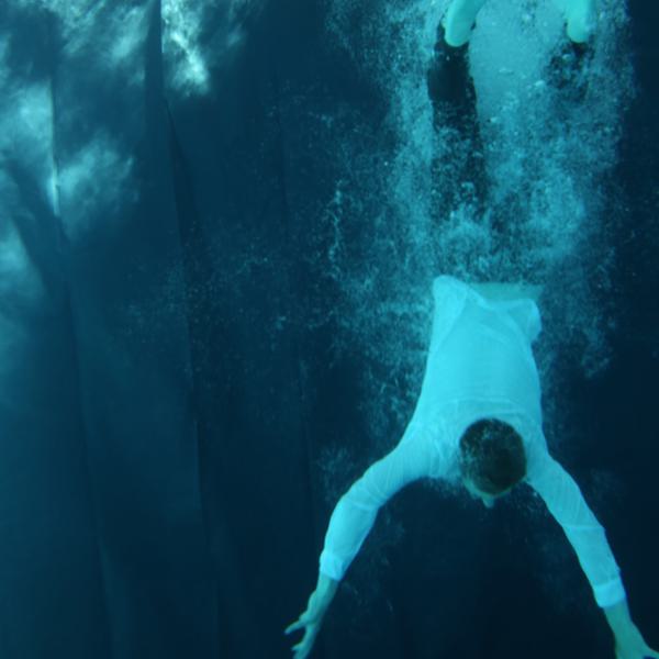 a man is plunging vertically into a pool, fully dressed with a whit shirt and black trousers.