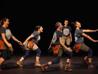 A group of young people in matching khaki shorts and bleached t shirts lunge across the stage in a line