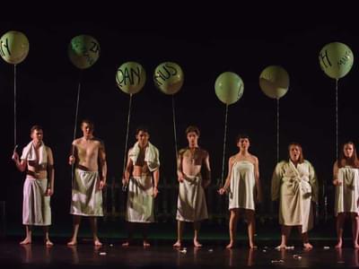 A group of young men and women stand in a line, dressed in towels holding balloons