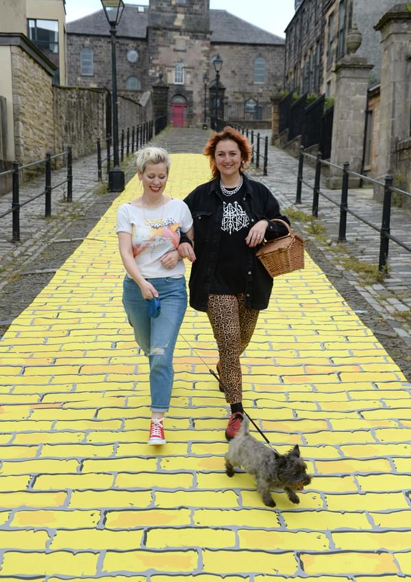A yellow brick road. 2 women walk down the centre with Toto.