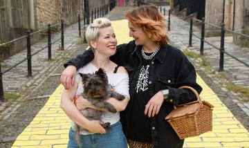 A yellow brick road. There are 2 women, one is holding Toto and one is holding a wicker picnic basket.