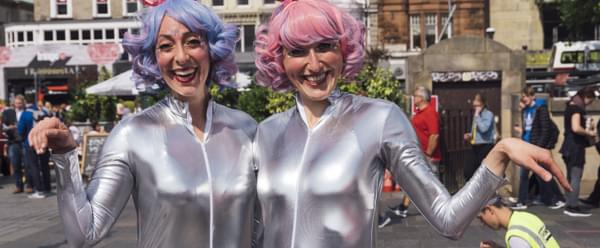 Photograph of two women in silver jump suits and pastel wigs