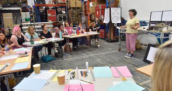 Workshop setting in a large studio with shelves of materials and equipment in the background. People sit in a semi circle of tables tables with coloured paper, pens, and cups, while the presenter stands at the front beside a screen and flip chart.