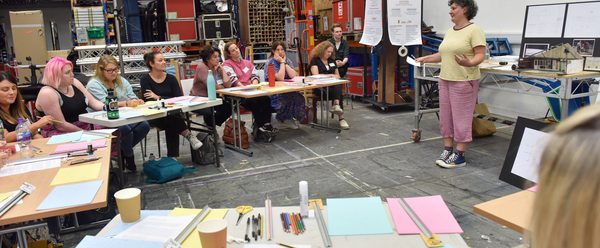 Workshop setting in a large studio with shelves of materials and equipment in the background. People sit in a semi circle of tables tables with coloured paper, pens, and cups, while the presenter stands at the front beside a screen and flip chart.