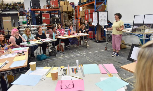 Workshop setting in a large studio with shelves of materials and equipment in the background. People sit in a semi circle of tables tables with coloured paper, pens, and cups, while the presenter stands at the front beside a screen and flip chart.