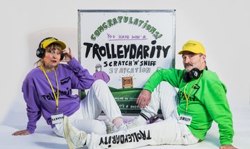 Two people are sat on either side of a poster for Trolleydarity. They are wearing branded tracksuits, one with a purple jumper and the other with a green jumper. They are both wearing yellow hats and headphones, one with a lanyard that says 'scratch' and the other that says 'sniff'.