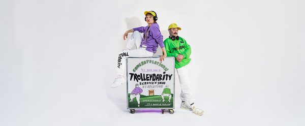 A trolley sits in front of a white background, the words "Trolleydarity" are on the trolley. Shona wears a purple top and sits atop the trolley. Alan stands beside the trolley. The pair look very cool.