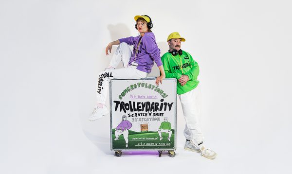 A trolley sits in front of a white background, the words "Trolleydarity" are on the trolley. Shona wears a purple top and sits atop the trolley. Alan stands beside the trolley. The pair look very cool.