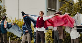 Group of performers stand outdoors in a line in front of a building and greenery. The central performer in a red vest extends both arms outward, while the rest of the performers behind and on either side hold flowing blue, pink, and patterned cloths, creating a wing-like visual effect.