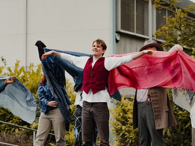 Group of performers stand outdoors in a line in front of a building and greenery. The central performer in a red vest extends both arms outward, while the rest of the performers behind and on either side hold flowing blue, pink, and patterned cloths, creating a wing-like visual effect.