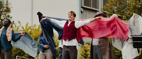 Group of performers stand outdoors in a line in front of a building and greenery. The central performer in a red vest extends both arms outward, while the rest of the performers behind and on either side hold flowing blue, pink, and patterned cloths, creating a wing-like visual effect.