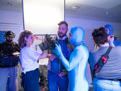 Film crew members mill about. A man in a body suit looks at footage from a camera
