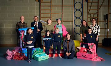 A group photo in a gym features adults and children with colorful props, including ribbons and suitcases. The mood is cheerful and creative, reflecting a playful atmosphere.
