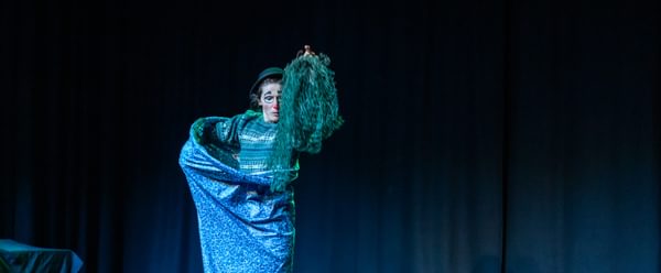 A performer dressed as a clown is on-stage. The lighting is a bluish-green.