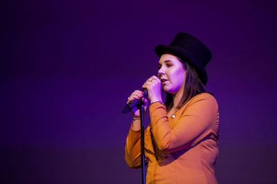 Woman in orange shirt and black top hat speaking into microphone