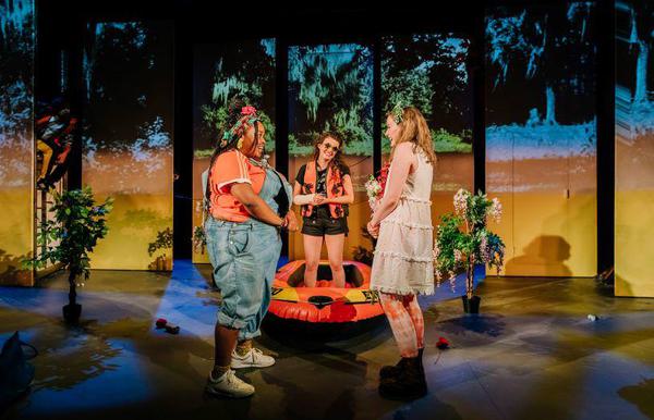 Three women on a tropical-looking set, one stands inside a rubber dinghy