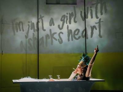 'I'm just a girl with a sharks heart' is written on the wall, a woman sits in the bath smoking and bearing her middle finger