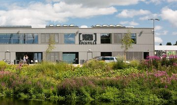 A grey two-storey building with trees and a canal in front of it