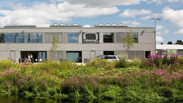 A grey two-storey building with trees and a canal in front of it
