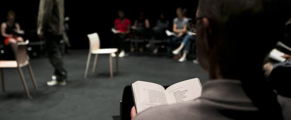 a person holding an open script or book in a dark rehearsal space, with a group seated around the rehearsal space in the background and two chairs on the floor under stage lighting.