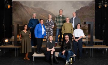 A group of people stand on platforms, in front of a painted backdrop of the Scottish Highlands, with large black brackets on either side and stage lights.