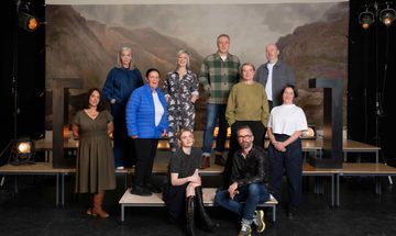 A group of people stand on platforms, in front of a painted backdrop of the Scottish Highlands, with large black brackets on either side and stage lights.