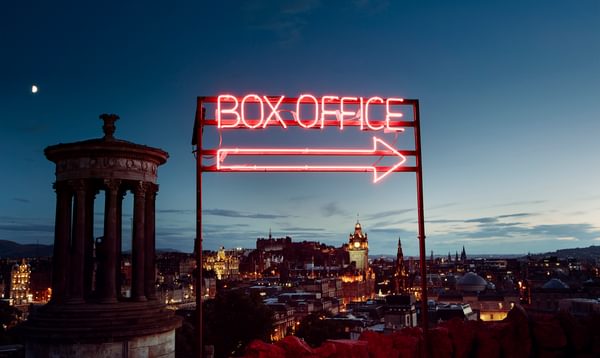 A neon box office sign with a view of Edinburgh at dusk behind.