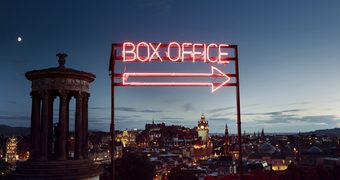 A neon box office sign pointing to the right sits on Calton Hill in front of a view of Edinburgh at dusk.