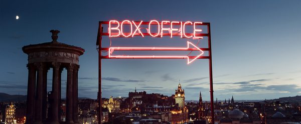 A neon box office sign pointing to the right sits on Calton Hill in front of a view of Edinburgh at dusk.