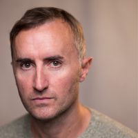Actor Ewan Donald wears a grey jumper and looks seriously at the camera, his eyes are bright